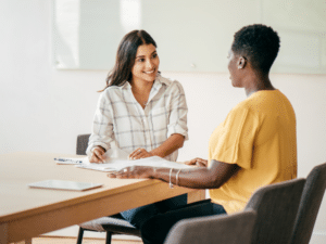 two women with one talking to the other about an apprentice career guide, they are very close and smiley, they are both sat next to each other at a wooden desk, dressed in casual clothes.